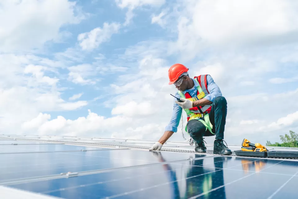 Worker wearing orange hard hat and vest over light blue long-sleeved shirt and jeans inspects rooftop solar panel